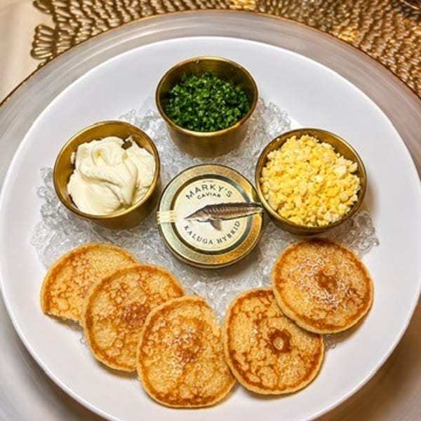 Plate of golden blinis arranged around caviar, crème fraîche, chopped egg, and herbs in small bowls.