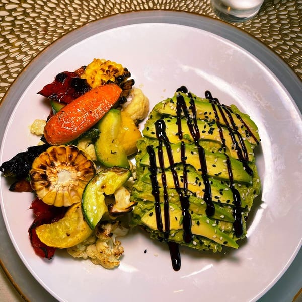 Plate of grilled vegetables and corn with sliced avocado drizzled in dark sauce and sprinkled with sesame seeds.