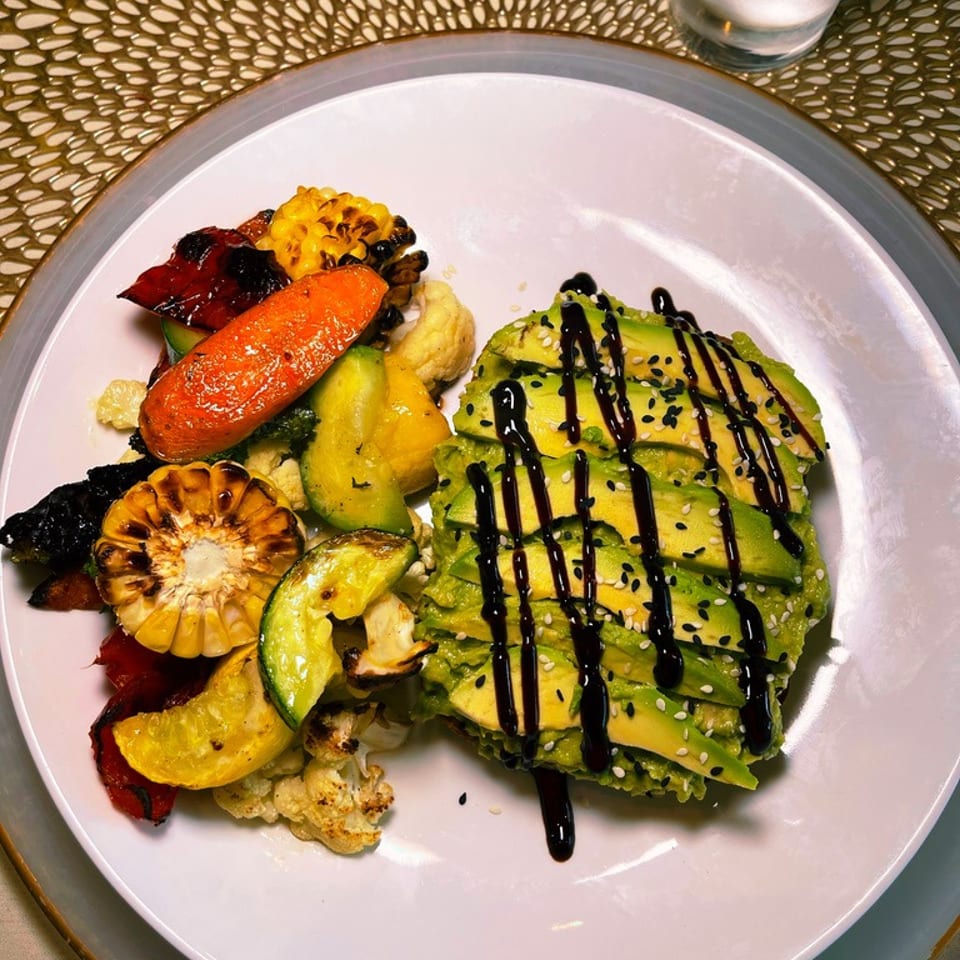 Plate of grilled vegetables and corn with sliced avocado drizzled in dark sauce and sprinkled with sesame seeds.